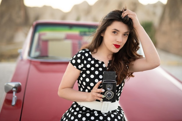 Hermosa chica en estilo retro posando cerca de un auto descapotable rojo vintage con una vieja cámara de cine en las manos