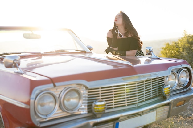 Foto hermosa chica en estilo retro posando cerca de un coche descapotable rojo vintage