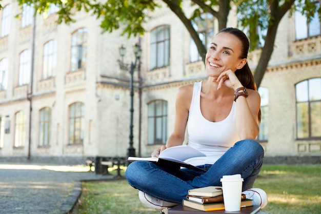 Hermosa chica estudiante morena feliz en ropa casual está sentada en el banco con libros y aprendiendo sobre antecedentes universitarios