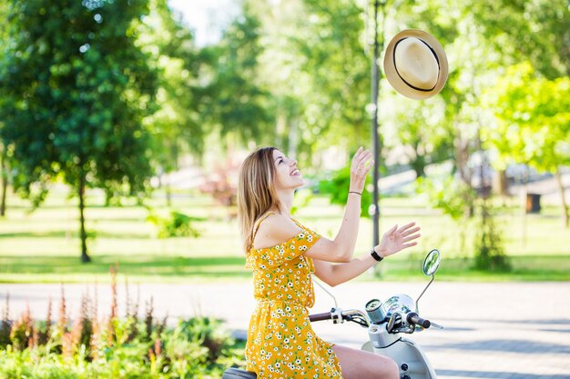 Hermosa chica europea con un vestido amarillo con un scooter retro en el parque de la mañana