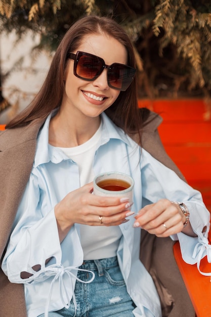 Hermosa chica feliz con una linda sonrisa con gafas de sol vintage en ropa de moda con un abrigo y una camisa se sienta en la calle y bebe té en un soleado día de primavera