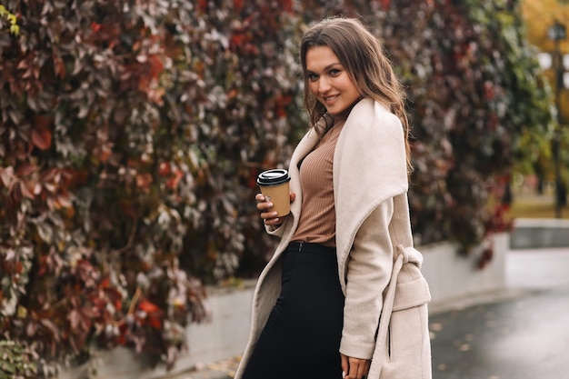 Una hermosa chica feliz con ropa casual está caminando en el parque al aire libre de otoño bebiendo café