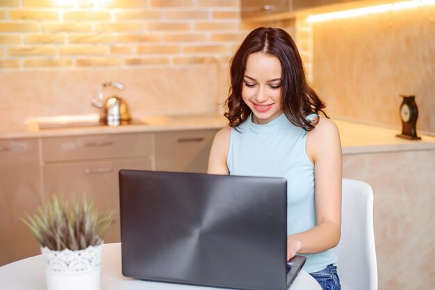 Foto hermosa chica feliz trabajando en la computadora sentada en la mesa en casa. apasionada por el trabajo. ropa elegante y de negocios. mira la pantalla del portátil.