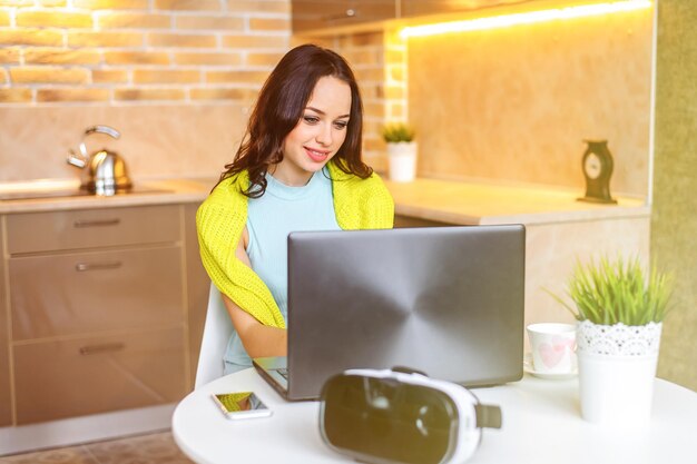 Hermosa chica feliz trabajando en la computadora sentada en la mesa en casa. Apasionada por el trabajo. Ropa elegante y de negocios. Mira la pantalla del portátil.