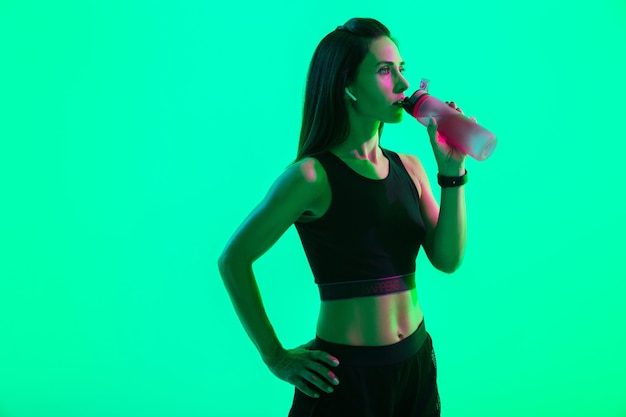 Hermosa chica de fitness joven confiada que se encuentran aisladas sobre la pared de neón verde, usando auriculares inalámbricos, bebiendo agua de una botella después del entrenamiento