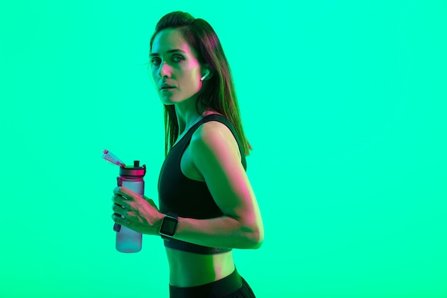 Hermosa chica de fitness joven confiada que se encuentran aisladas sobre la pared de neón verde, usando auriculares inalámbricos, sosteniendo una botella de agua después del entrenamiento
