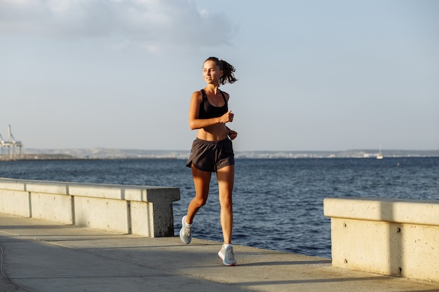Hermosa chica fitness en pantalones cortos y top deportivo se ejecuta en el paseo marítimo junto al mar durante la puesta de sol
