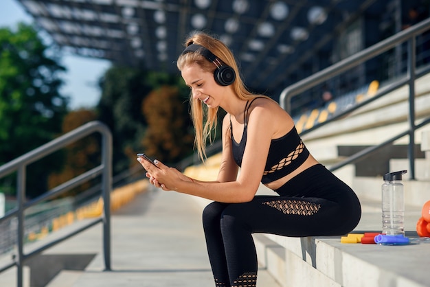 Una hermosa chica de fitness en ropa deportiva gris usa un teléfono inteligente y escucha música en el estadio después del entrenamiento.
