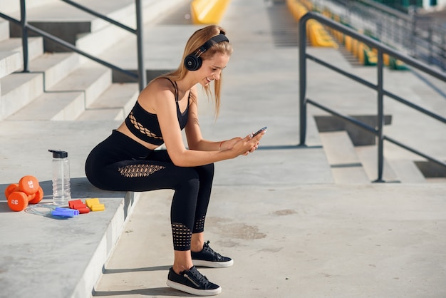 Una hermosa chica de fitness en ropa deportiva gris usa un teléfono inteligente y escucha música en el estadio después del entrenamiento.