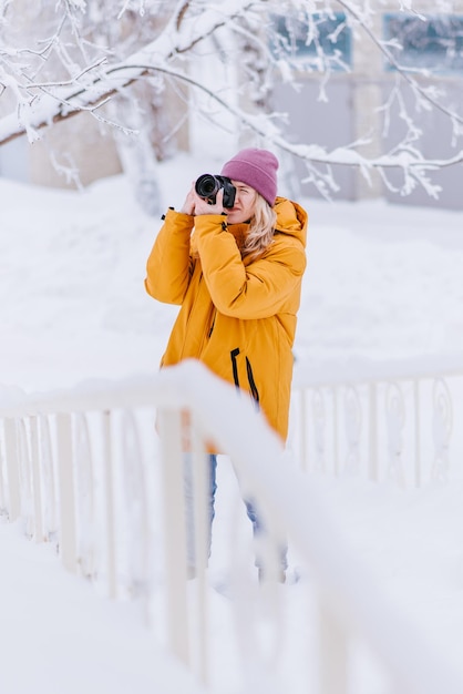 Hermosa chica en un fotógrafo de chaqueta amarilla toma fotos de nieve en un parque de invierno