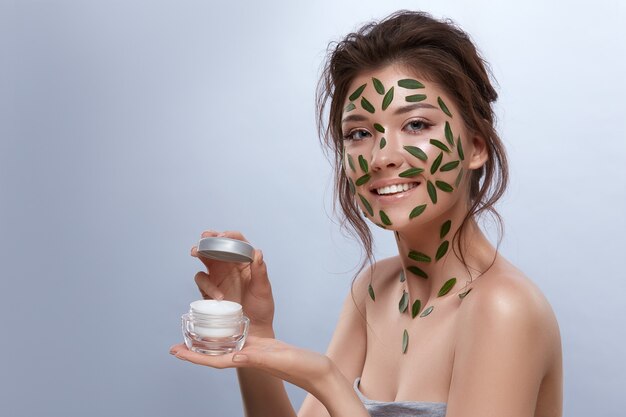 Hermosa chica fresca con maquillaje natural y hojas verdes en el cuerpo con crema y mirando a la cámara