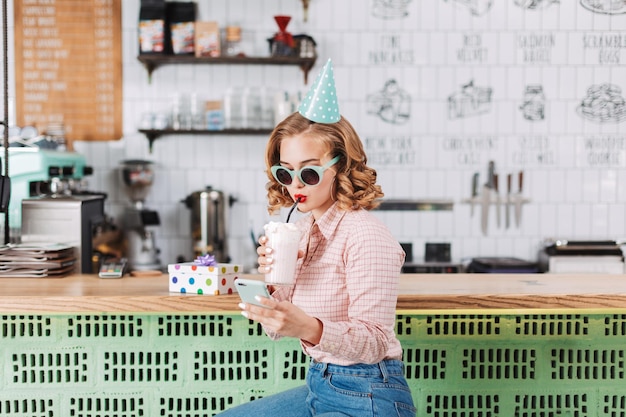 Hermosa chica con gafas de sol y gorra de cumpleaños sentada en el mostrador del bar y bebiendo batido mientras usa su celular en el café