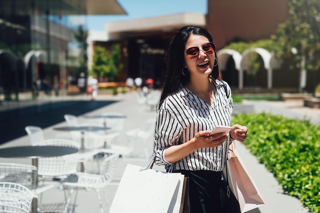 Foto hermosa chica con gafas de sol sosteniendo bolsas de compras usando un teléfono inteligente y sonriendo mientras está de pie al aire libre. mujer asiática riendo chateando en línea en el teléfono celular en un día soleado mientras camina por el centro comercial de stanford