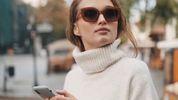 Hermosa chica con gafas de sol vestida con un suéter blanco acogedor que se ve elegante usando un teléfono inteligente en la calle de la ciudad