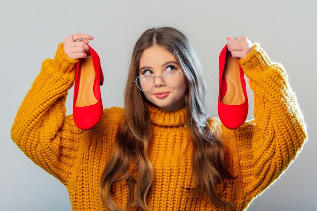 Hermosa chica con gafas y suéter amarillo con tacones rojos sobre fondo blanco.