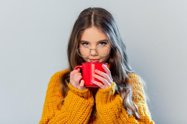 Hermosa chica con gafas y suéter amarillo con taza roja sobre fondo blanco.