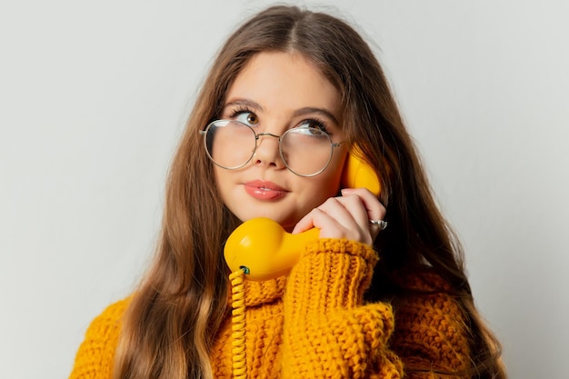 Hermosa chica con gafas y suéter amarillo con teléfono amarillo sobre fondo blanco.