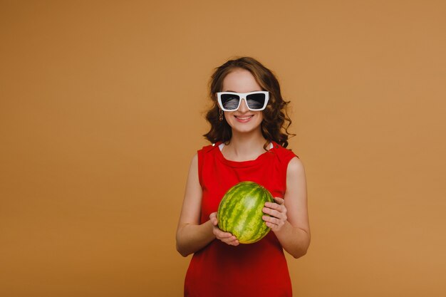 Una hermosa chica con gafas y un vestido rojo sostiene una sandía en sus manos.