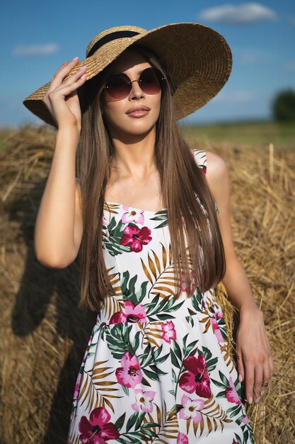 Hermosa chica glamorosa con un vestido y un sombrero de paja en un campo cerca de un pajar