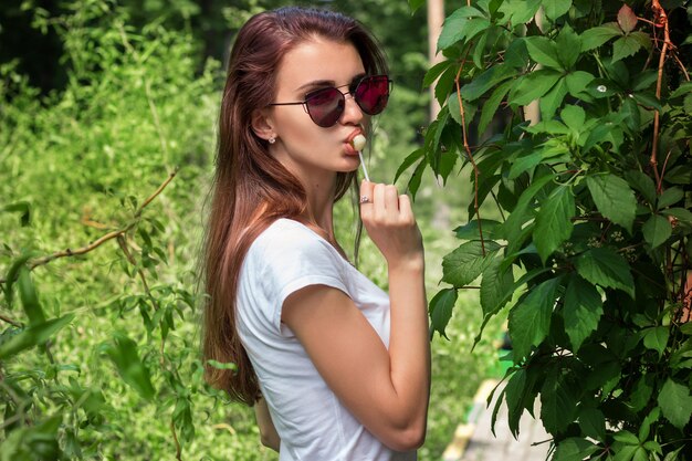 Hermosa chica glamour en gafas brillantes lame una piruleta y mira a la cámara de cerca