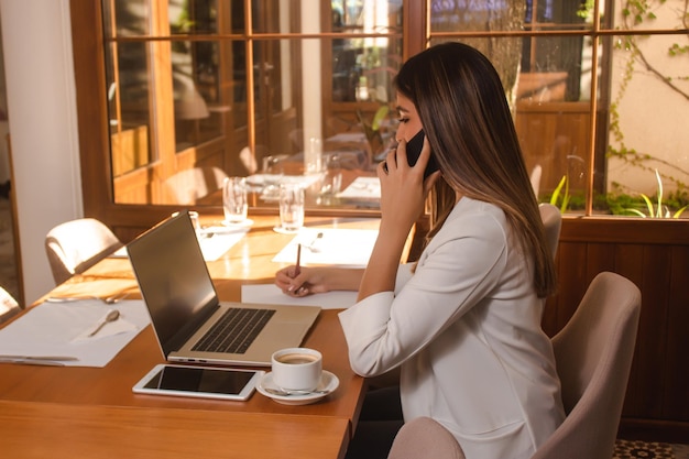 Hermosa chica hablando por teléfono mientras toma notas en un papel en el restaurante