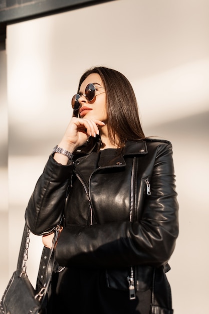 Hermosa chica hipster con estilo con gafas de sol vintage en una chaqueta de cuero de moda con un bolso negro de moda camina en la ciudad al atardecer