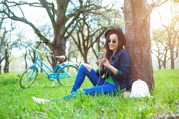 Foto hermosa chica hipster con estilo sonriente con sombrero y gafas de sol está sentada en un parque debajo de un árbol con un teléfono en las manos y escuchando música en un fondo de bicicleta retro