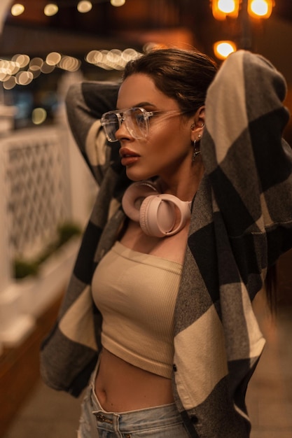 Hermosa chica hipster de moda con gafas y auriculares rosas con ropa informal de moda con un top y una camisa a cuadros camina por la ciudad por la noche