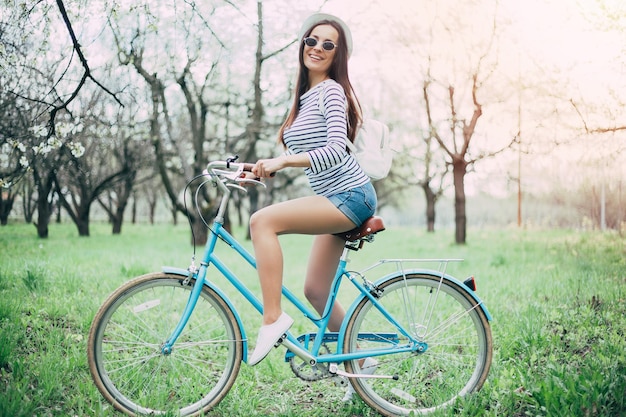 Hermosa chica hipster moderna con gafas y un sombrero blanco se sienta en una bicicleta a la luz del sol y mira a la cámara