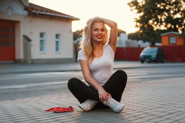 Hermosa chica hipster sentada en el asfalto cerca de una carretera al atardecer