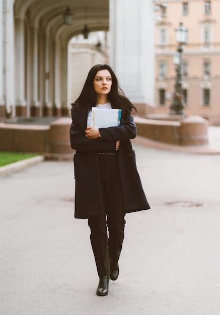 Hermosa chica inteligente seria morena estudiante sosteniendo cuadernos y libros de texto, va caminando