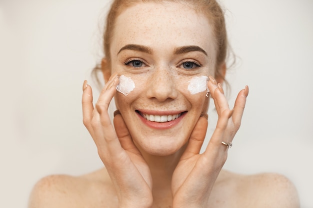 Hermosa chica de jengibre aplicando una crema especial en su rostro pecoso mientras posa en una pared beige