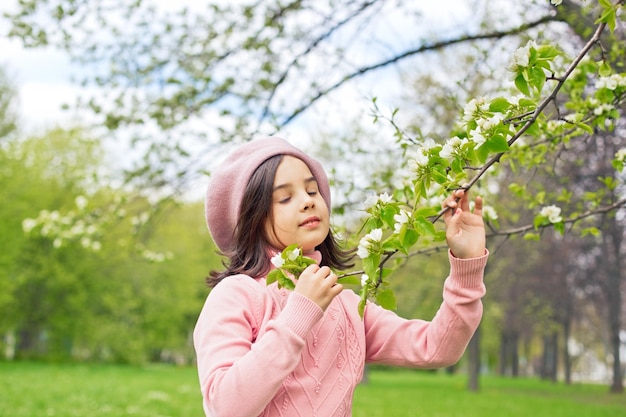 Una hermosa chica con un jersey rosa sostiene una rama de un manzano blanco floreciente en el parque durante el día