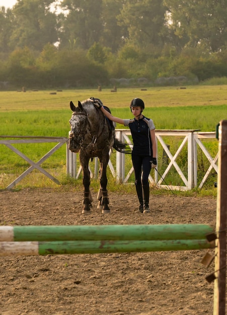 Hermosa chica jockey de pie junto a su caballo con uniforme especial en un cielo y fondo de campo verde