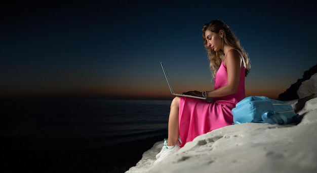 Una hermosa chica con una laptop con un vestido rosa junto al mar entre las rocas