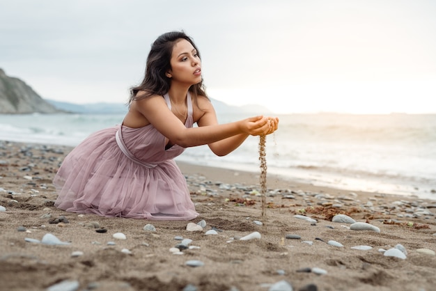 Hermosa chica latina con el palo largo negro posando de rodillas en el suelo y jugando con la arena en sus manos en una hermosa puesta de sol en la playa vestida con un vestido de ballet rosa