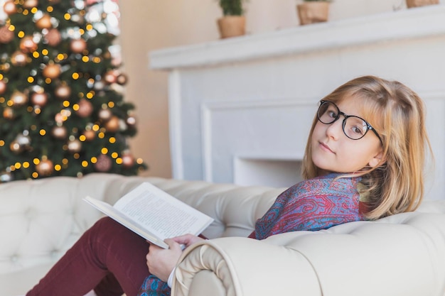Hermosa chica leyendo un libro en el sofá cerca del árbol de navidad