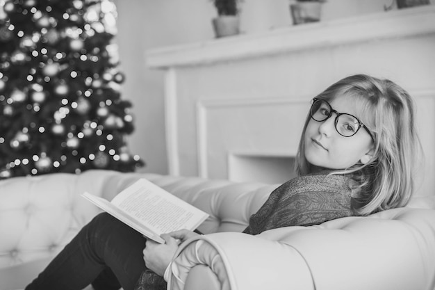 Hermosa chica leyendo un libro en el sofá cerca del árbol de navidad