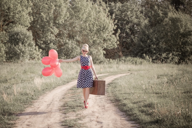 Hermosa chica con maleta y globos rojos en el campo