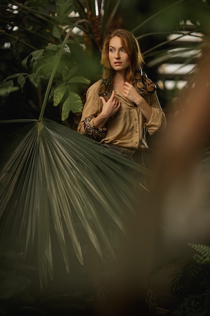 Una hermosa chica con maquillaje natural y cabello rojo se encuentra en la jungla entre plantas exóticas con serpiente.