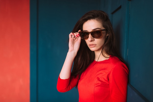 Hermosa chica de moda con cabello largo oscuro, apariencia española en gafas de sol y elegante vestido rojo posando en la pared azul rojo.