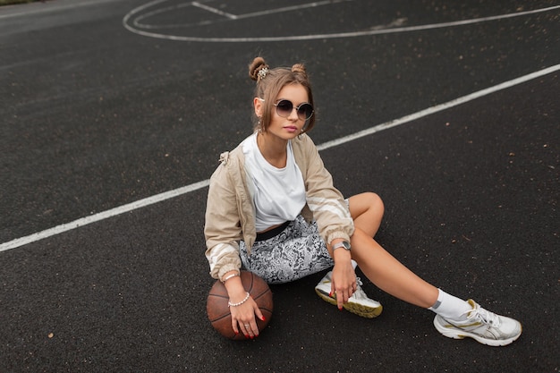 Hermosa chica de moda con peinado y gafas de sol en una chaqueta deportiva de moda, pantalones blancos, camiseta y zapatillas con una pelota sentada en la calle en el pavimento