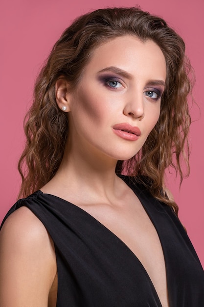 Hermosa chica de moda con pelo largo y rizado con un vestido negro en el estudio sobre un fondo rosa.