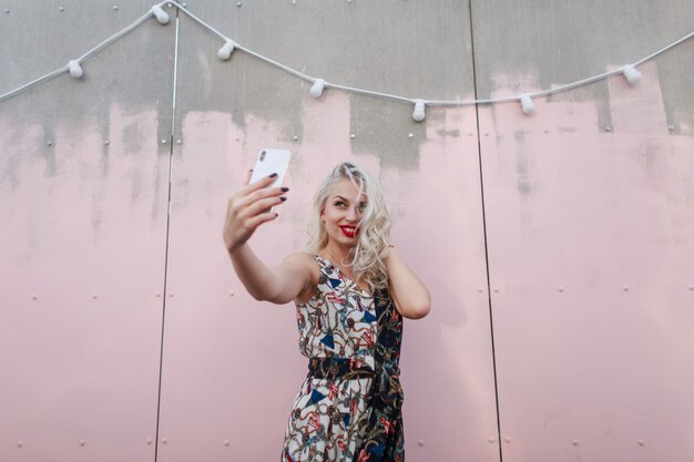 Hermosa chica de moda con un vestido de moda hace selfie en el teléfono cerca de la pared rosa en una fiesta