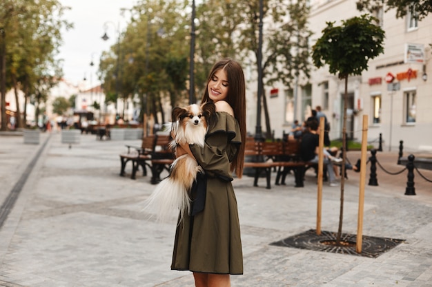 Hermosa chica modelo con amplia sonrisa en vestido corto posando con pequeño lindo papillon perro en sus manos al aire libre en el centro de la ciudad vieja. Moda de verano