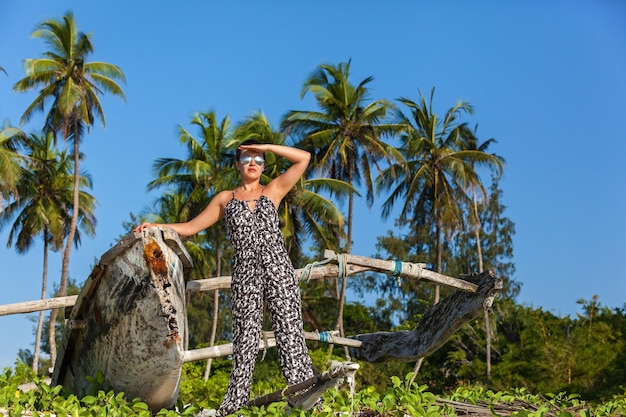 Hermosa chica en monos y gafas de sol posando en la playa con palmeras altas y arena blanca