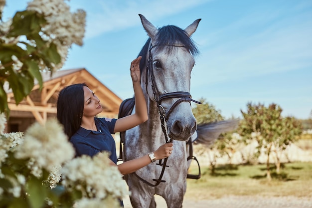 Hermosa chica morena acariciando su caballo gris cerca de arbustos de lilas en el jardín.