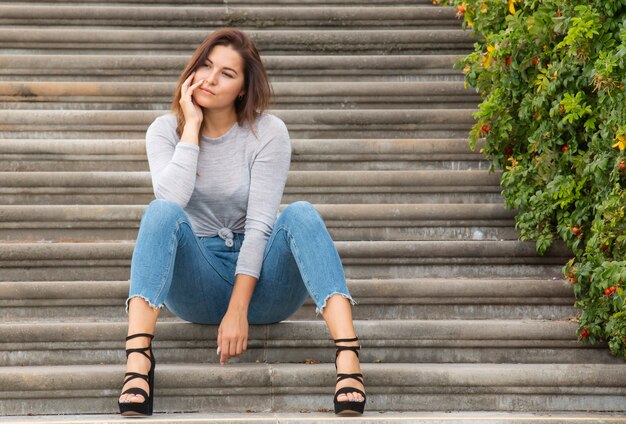 Hermosa chica morena al aire libre sentado en pasos concretos, arbustos de rosa mosqueta