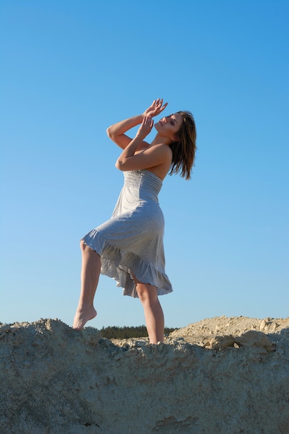 Hermosa chica morena bailando y mirando al sol. Chica con un vestido blanco en el desierto contra el cielo azul.