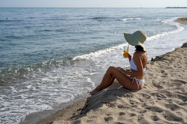 hermosa chica morena bronceada en un traje de baño blanco bebe de un vaso de jugo de naranja sentada en la arena en la playa junto al mar